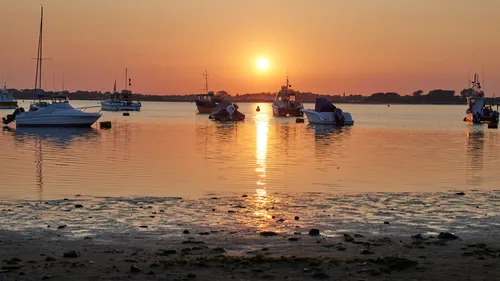 This landscape photograph captures an evening sunset over Christchurch Harbour in Dorset, United Kingdom. The warm light of the sun, low in the sky, reflects across the tranquil water, marking a late spring day. Several boats are anchored near the beach, emphasising the harbour’s role in local transport and maritime activity. The image features the natural beauty of Christchurch, with the boats silhouetted against the colourful sky and the shoreline in the foreground.